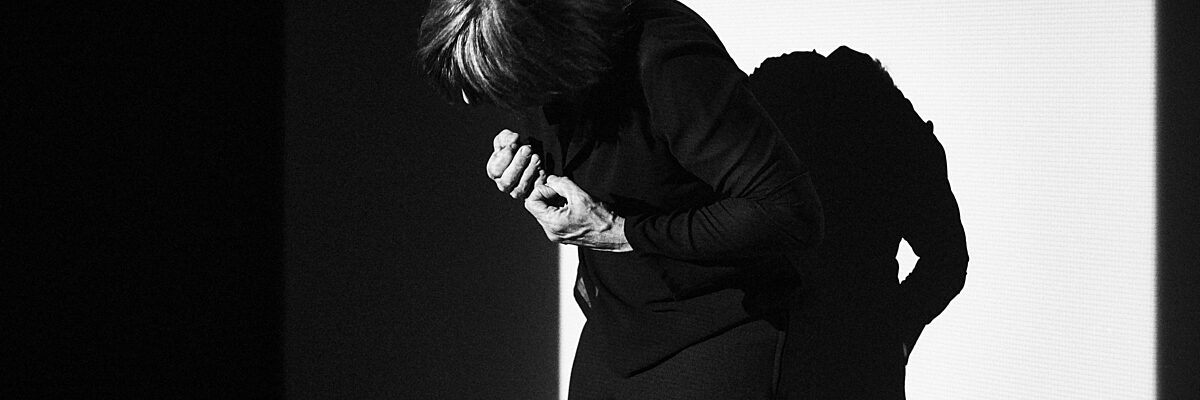 Een danser staat voorovergeboven met de rug naar een muur, het gezicht onherkenbaar, de handen in vuisten voor de borst geklemd.  

A dancer stands with the back against a wall, bending forward, face hidden, hands in clenched fists in front of the person.